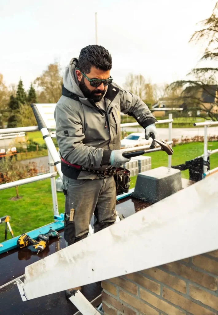Dakdekker van Astrodak werkt met een hamer aan een schoorsteen op een steiger tijdens dakwerkzaamheden
