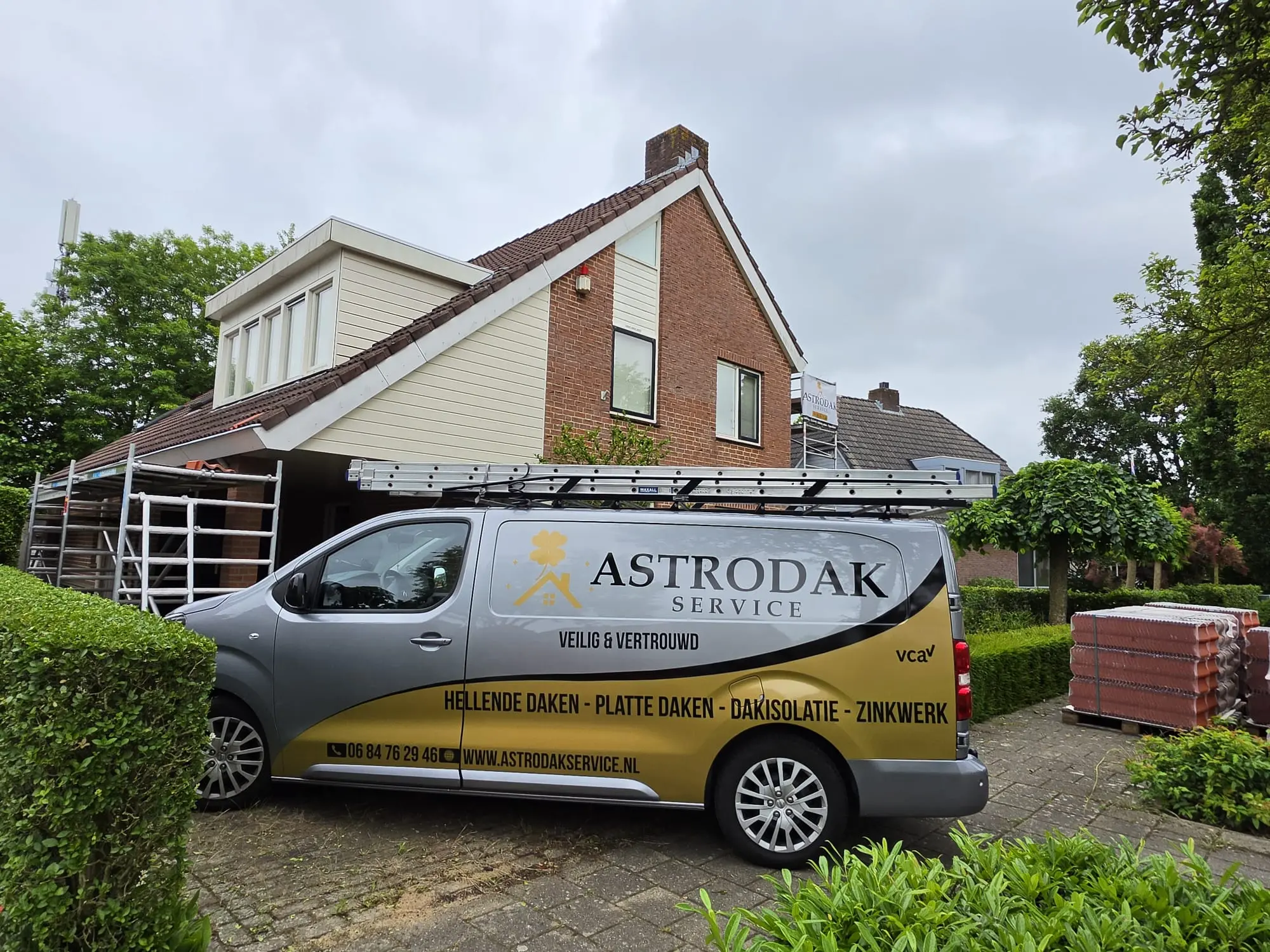 Bedrijfsbus van Astrodak Service geparkeerd bij een woning met dakkapel, met steiger en dakpannen klaar voor dakwerkzaamheden
