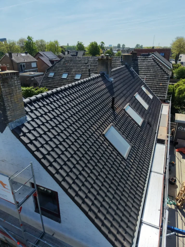 Hellend dak met zwarte dakpannen en dakramen onder een blauwe lucht, met omliggende woningen