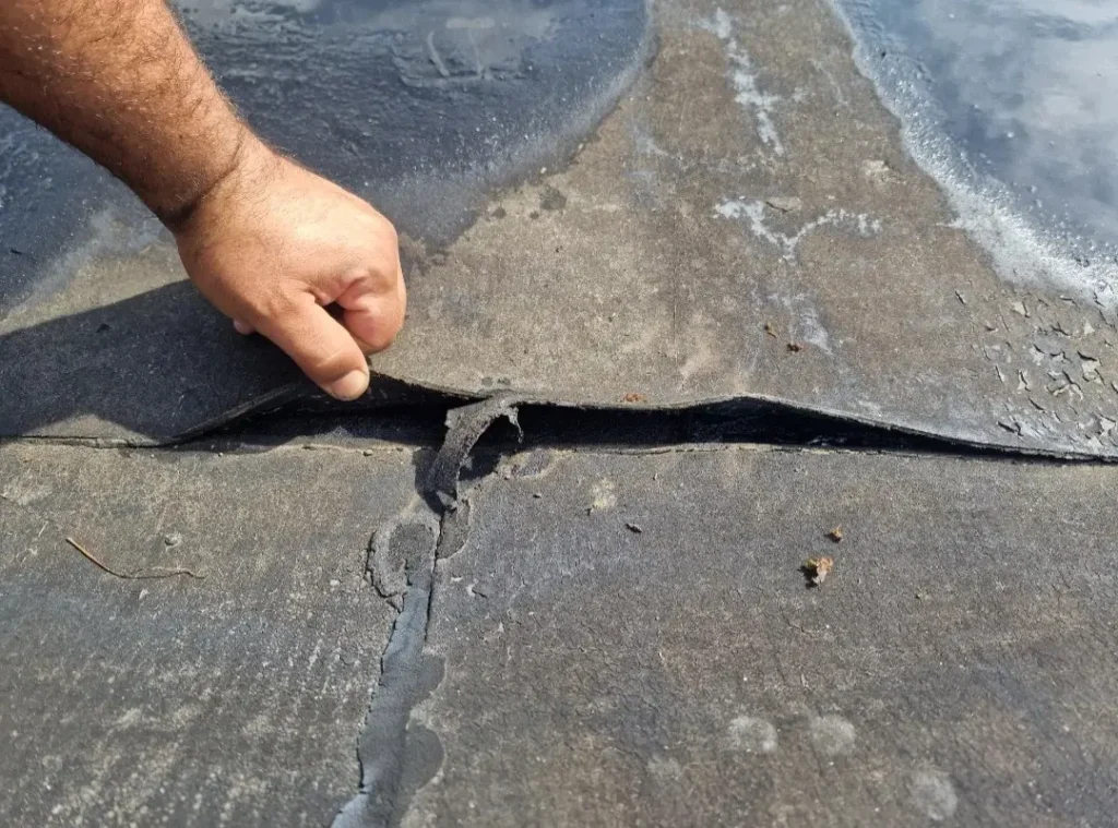 Hand toont gescheurde en beschadigde bitumen dakbedekking op een plat dak met lekkage